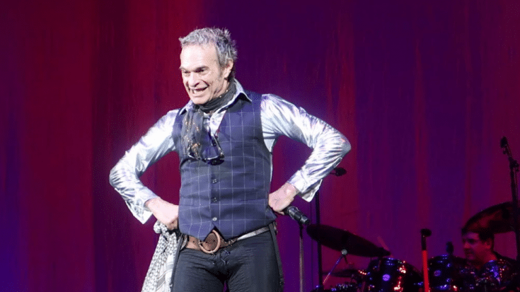 David Lee Roth in a stylish outfit performing on stage with a live band in the background.
