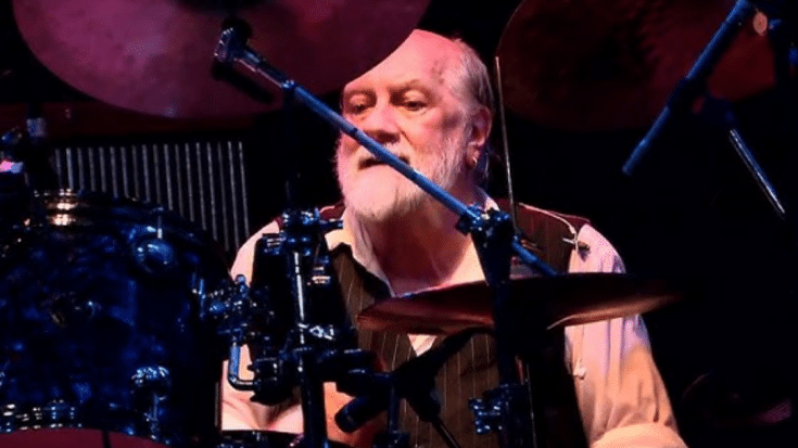 Mick Fleetwood with a white beard, wearing a vest, seated at a drum kit during a live performance.