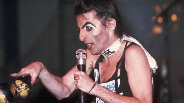 Alice Cooper performing on stage with dramatic makeup, holding a microphone with a skull design and shaking maracas.