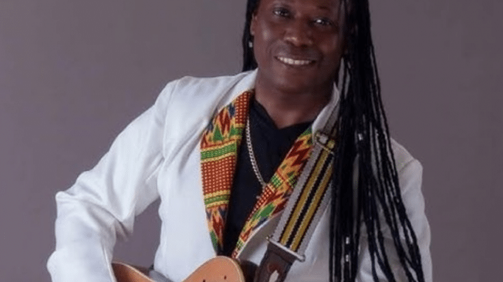Aurelio Martinez smiling while playing an acoustic-electric guitar, wearing a white blazer and a colorful patterned scarf.