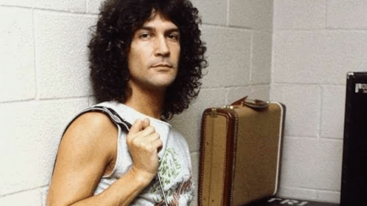 Billy Squier posing backstage with a suitcase and equipment case nearby.