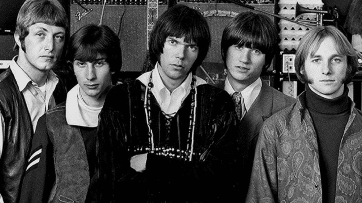 Buffalo Springfield members in a black-and-white setting, standing together with serious expressions, surrounded by amplifiers and musical equipment.