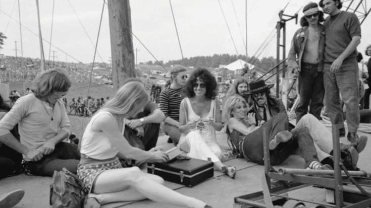 Jefferson Airplane members and friends relaxing backstage at an outdoor music festival, with Grace Slick smiling in the center and a large crowd visible in the background.