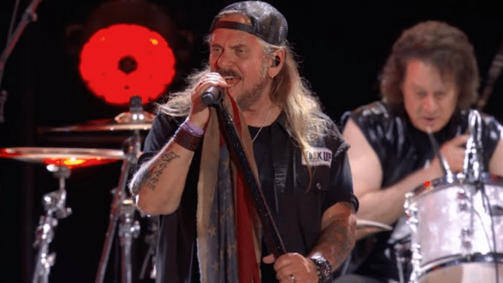 Lynyrd Skynyrd performing live, with the lead singer gripping a microphone stand draped in an American flag scarf while the drummer plays in the background.