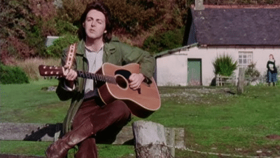 Paul McCartney sitting on a wooden fence, playing an acoustic guitar in a rural setting, with a small cottage in the background during the "Mull of Kintyre" music video.