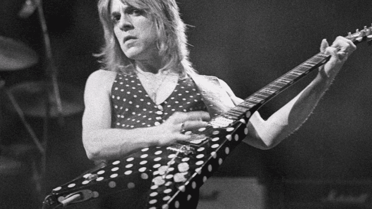 Randy Rhoads playing his signature polka-dot Flying V guitar on stage, wearing a sleeveless polka-dot shirt, with a focused expression.