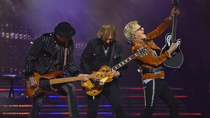REO Speedwagon performing live on stage, with three band members playing guitars under colorful stage lighting. The musicians are dressed in stylish outfits, with one guitarist raising his instrument in an energetic pose.