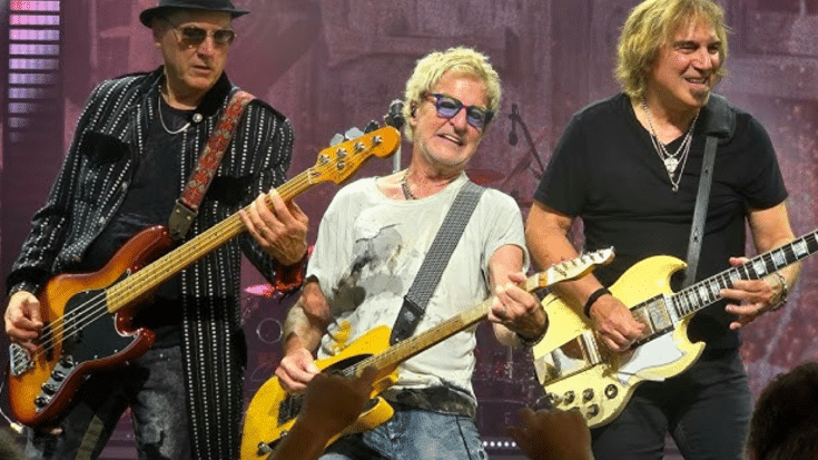 REO Speedwagon performing on stage, with three band members playing guitars and engaging with the audience. The musicians are dressed in casual and stylish outfits, smiling as they perform under bright stage lights.