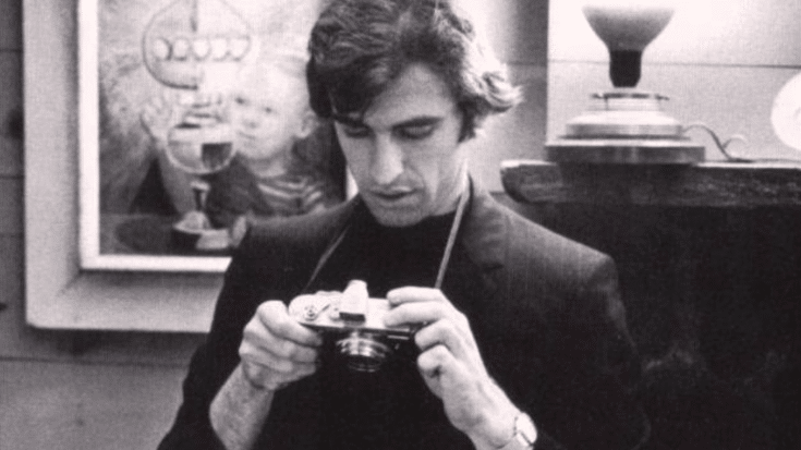 Black-and-white photo of Richard Manuel in a suit examining a camera, with a painting visible in the background.