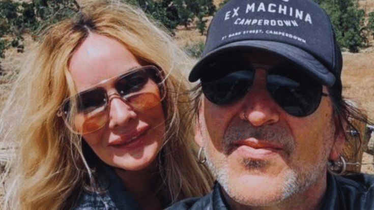 Taime Downe wearing a black cap and sunglasses, alongside Kimberly Burch, who has long blonde hair and is also wearing sunglasses, both outdoors with a natural landscape in the background.