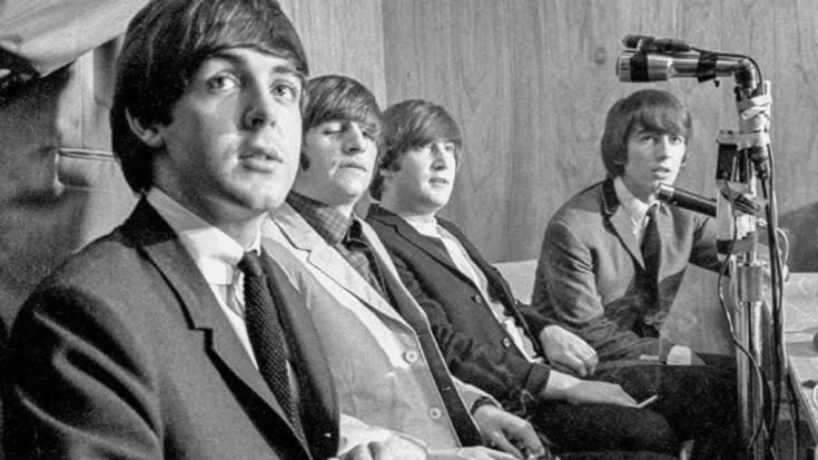 The Beatles at a press conference, with Paul McCartney, Ringo Starr, John Lennon, and George Harrison dressed in suits, seated together, and speaking into microphones.
