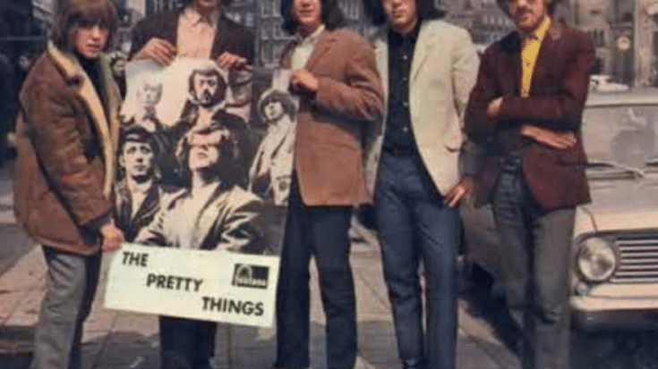 The Pretty Things standing on a city street, dressed in stylish 1960s attire, holding a large promotional poster of themselves.