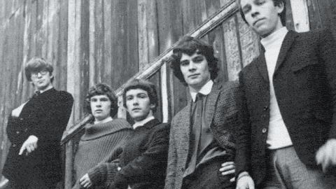 The Zombies standing against a wooden wall in the 1960s, dressed in classic mod-style clothing with a serious expression.