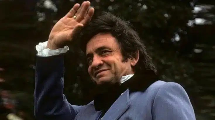 Johnny Cash smiling and waving in a light-colored suit, captured outdoors with trees in the background.