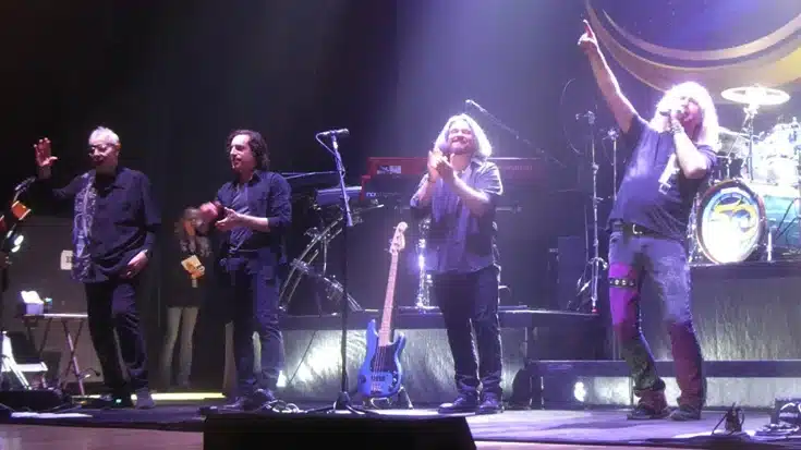 Members of Kansas take a bow on stage during a recent live performance, with instruments behind them and stage lights casting a purple hue.