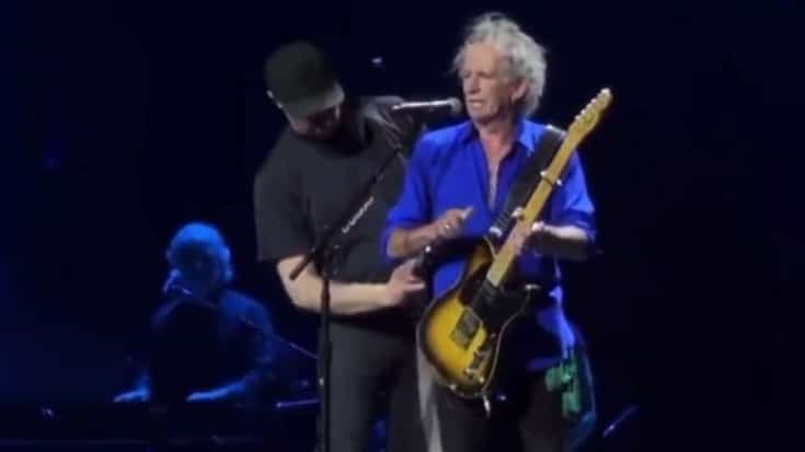 Keith Richards onstage mid-performance, being assisted by a crew member with his guitar, maintaining a cool and composed expression under the spotlight.