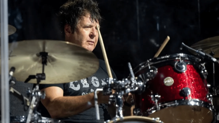 Clem Burke playing drums with focus and intensity, wearing a dark shirt behind a red sparkle drum kit on stage.