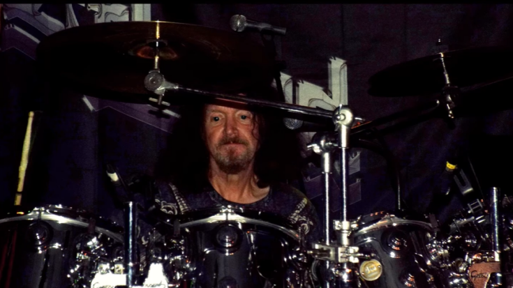 Les Binks behind a drum kit during a live heavy metal performance, surrounded by cymbals and stage lights