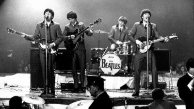 The Beatles performing live on stage in the 1960s, featuring Paul McCartney, George Harrison, Ringo Starr, and John Lennon in suits with their instruments.