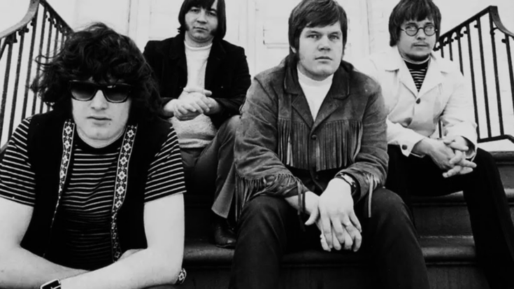 The Guess Who sit on outdoor steps, wearing 1960s-style outfits including striped shirts, fringe jackets, and sunglasses, with relaxed expressions.