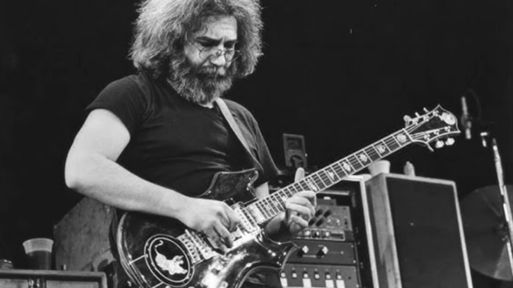 Jerry Garcia playing electric guitar on stage during a live Grateful Dead performance, wearing a black t-shirt and glasses