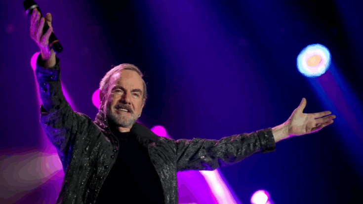 Neil Diamond performing live on stage with arms outstretched under purple concert lights