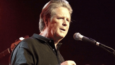 Brian Wilson performing live on stage in his later years, holding a microphone with a focused expression under stage lighting