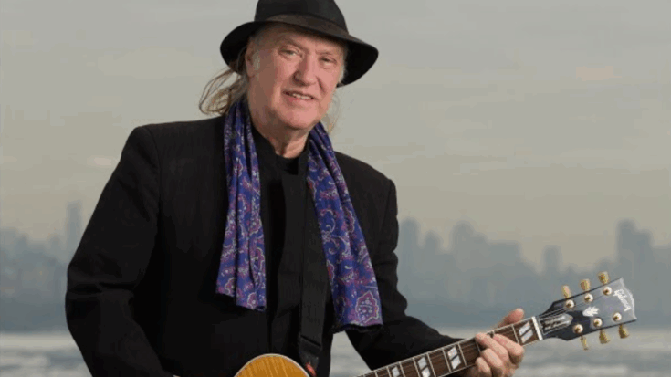Dave Davies wearing a black hat and paisley scarf while playing guitar outdoors with a hazy city skyline in the background.