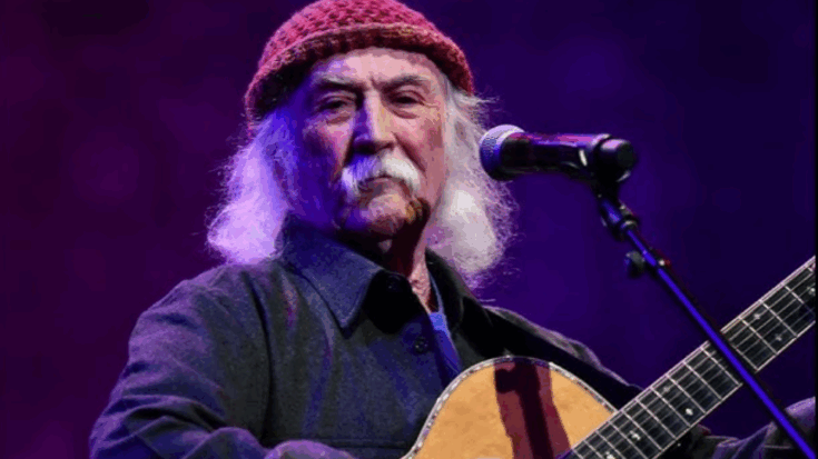 David Crosby performing live on stage with acoustic guitar and microphone under purple stage lighting