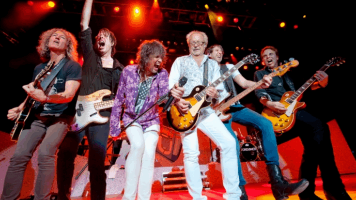 Foreigner performing on stage with vibrant lighting, all band members playing guitars or singing in an energetic pose during a live concert.