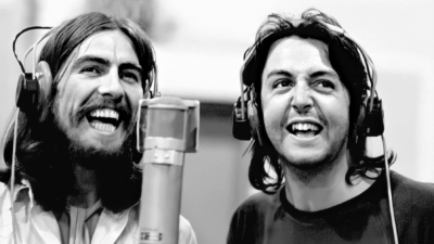 George Harrison and Paul McCartney smiling and wearing studio headphones while recording vocals together at a microphone