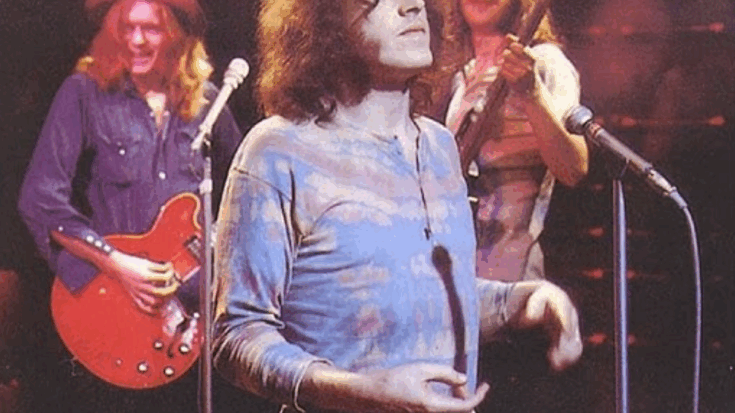 Joe Cocker stands at a microphone mid-performance, wearing a tie-dye shirt, with two guitarists smiling in the background under stage lights.