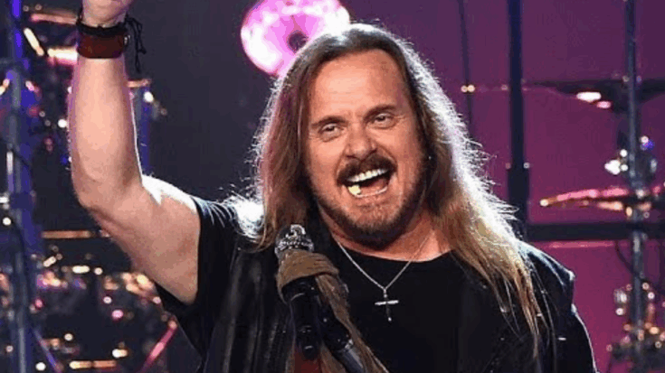 Johnny Van Zant performing on stage with one arm raised and a smile, wearing a black shirt, leather vest, and cross necklace, with drums and purple stage lights in the background.