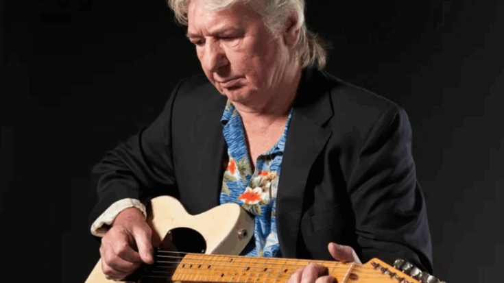 Mick Ralphs seated while playing a cream-colored electric guitar, wearing a floral shirt under a dark blazer against a black background