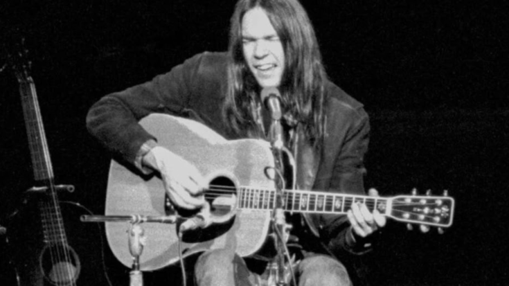 Neil Young sits on stage performing an acoustic set with a guitar in hand, eyes closed in mid-song, with another guitar resting nearby in the background.