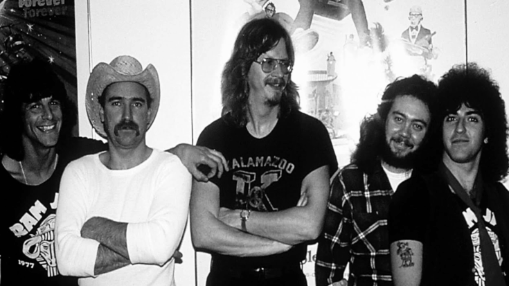 Ram Jam members standing together in a casual backstage setting, with 1970s-style clothing including graphic tees, a cowboy hat, and long hairstyles, appearing relaxed and smiling.