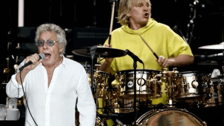 Roger Daltrey sings into a microphone in a white shirt while Zak Starkey plays drums in a bright yellow hoodie during a live performance.