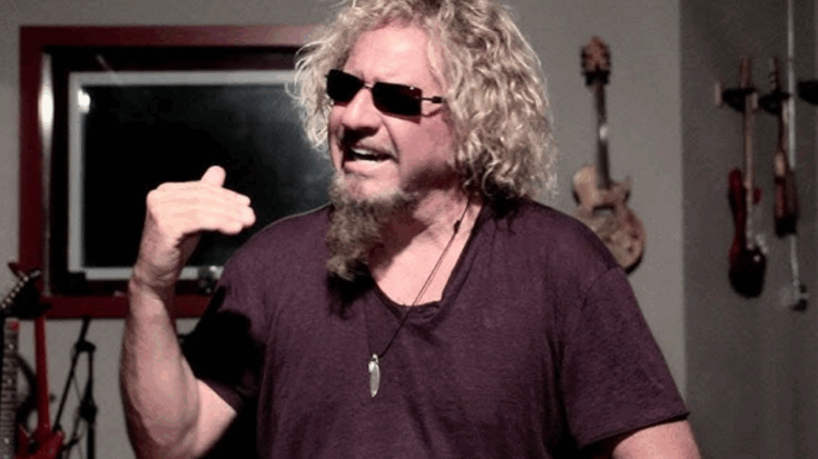 Sammy Hagar wearing sunglasses and a dark shirt, gesturing with one hand while speaking in a room with guitars hanging on the wall.