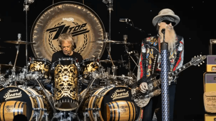Frank Beard plays an elaborate skull-themed drum kit with ZZ Top’s logo while Billy Gibbons performs beside him wearing a colorful jacket, sunglasses, and hat during a live concert.
