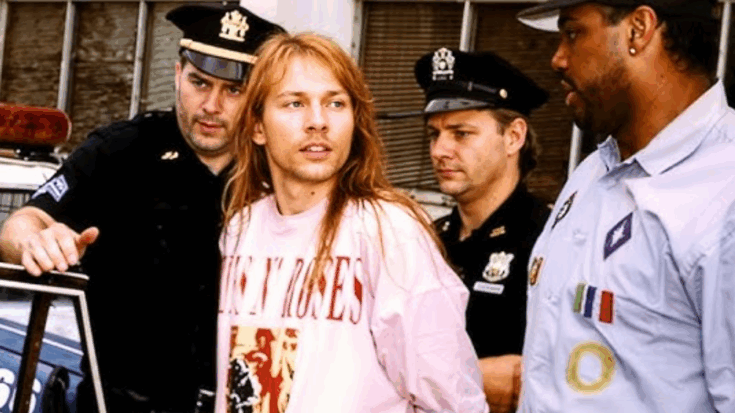 Axl Rose being escorted by police officers while wearing a Guns N' Roses sweatshirt, looking off to the side during an arrest scene.