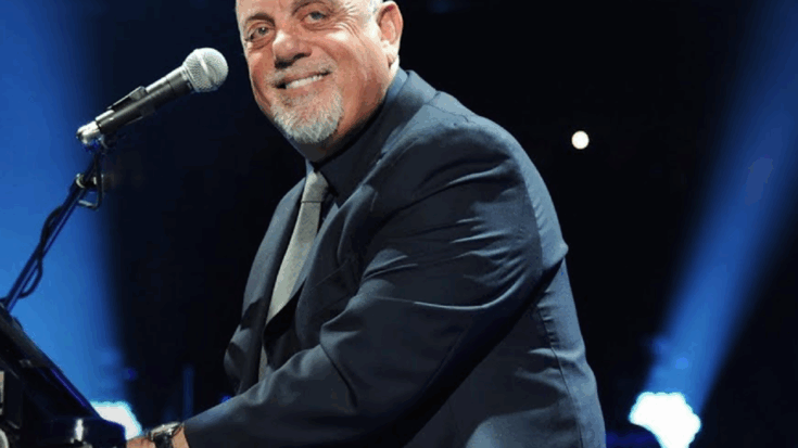 Billy Joel smiling while performing at a piano during a live concert, dressed in a dark suit with a microphone in front of him.