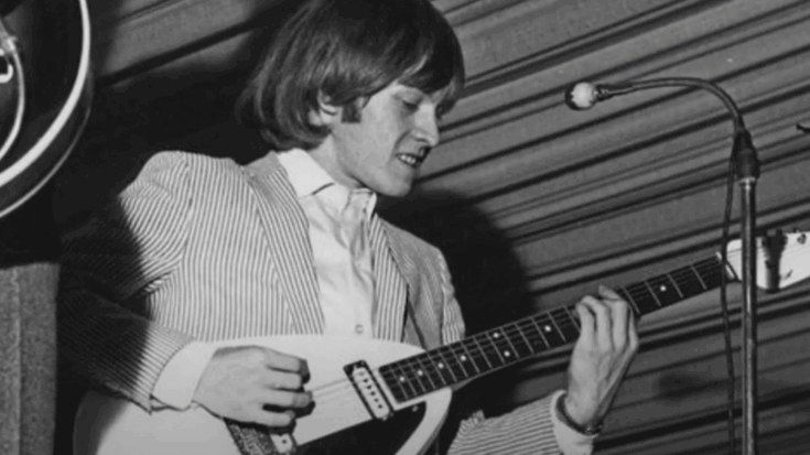 Brian Jones playing guitar onstage, wearing a striped jacket and focusing on his fretwork during a live performance.