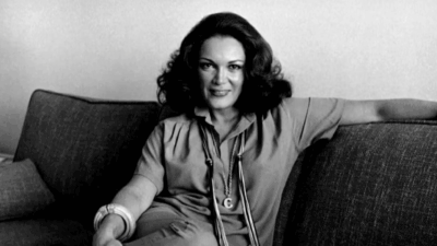 Connie Francis sitting on a couch, wearing a short-sleeve blouse and trousers, with a long necklace featuring a large "C" pendant, smiling confidently with her arm resting along the back of the sofa.