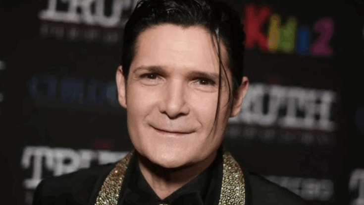 Corey Feldman wearing a black outfit with a gold-sequined collar and styled hair, standing in front of a dark event backdrop.