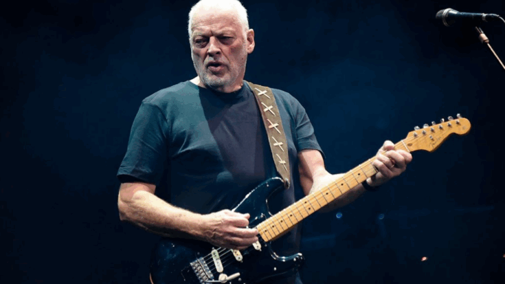 David Gilmour playing a black electric guitar on stage under dark lighting, wearing a black t-shirt with a brown strap over his shoulder.