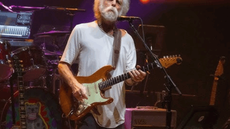 Dead and Company performing live with guitarist in a white shirt playing a sunburst Fender Stratocaster on a dimly lit stage