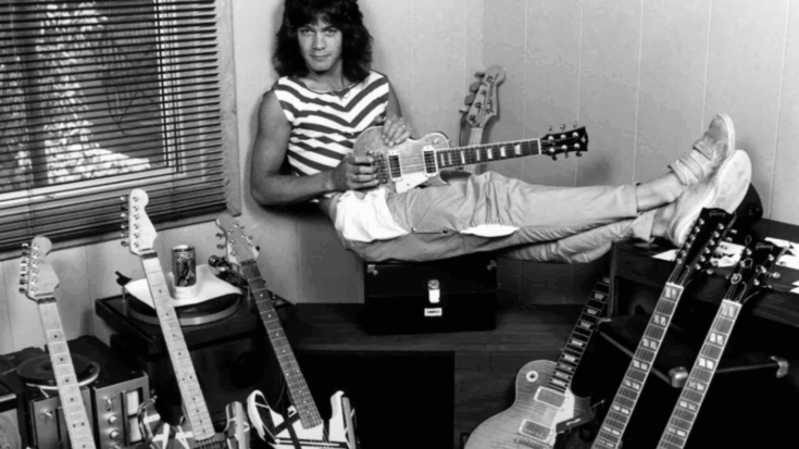 Eddie Van Halen lounging casually with a guitar in hand, surrounded by various electric guitars in a room with a window and blinds.