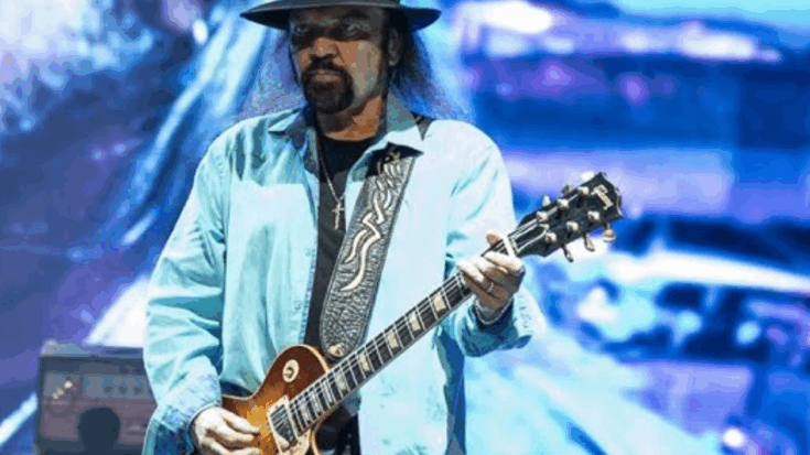 Gary Rossington wearing a light blue shirt and black hat while playing a Les Paul guitar on stage under blue stage lighting