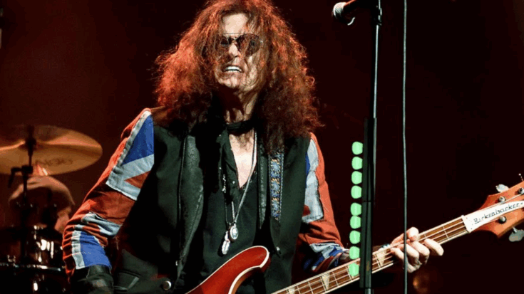 Glenn Hughes performing live on stage wearing a Union Jack jacket while playing a Rickenbacker bass guitar under red lighting.