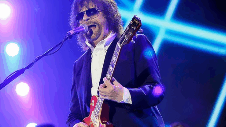 Jeff Lynne performing live on stage wearing sunglasses and playing a red electric guitar under colorful stage lights.